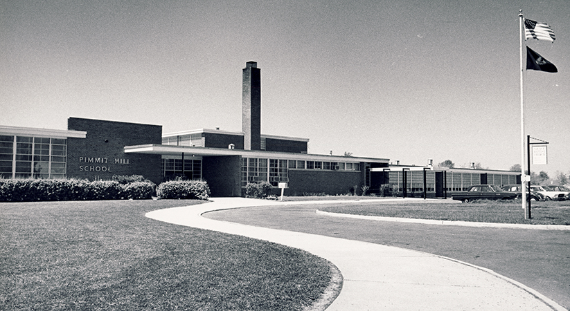 Black and white photograph of the main entrance of Pimmit Hills Elementary School taken in the late 1960s. The building is a one story structure with a tall chimney. There are banks of windows with white trim. The main entrance is located beneath an awning. The sidewalk snakes its way from the vantage point of the camera toward the building. A flag pole and several parked cars are visible to the right. 
