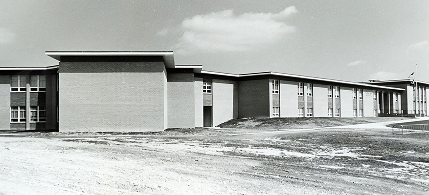 Black and white photograph of Westgate Elementary School taken in the late 1960s shortly after construction of the school had been completed. The school grounds are still bare-earth. The building's main entrance faces east and it appears this photo was taken before noon because of the low angle of the shadows. The building has a brick exterior and relatively few windows compared to other schools of the time. It's shape is different, too. It has an angled wing on the left (south) side, and is two stories tall. This style appears unique in all of Fairfax County Public Schools. 