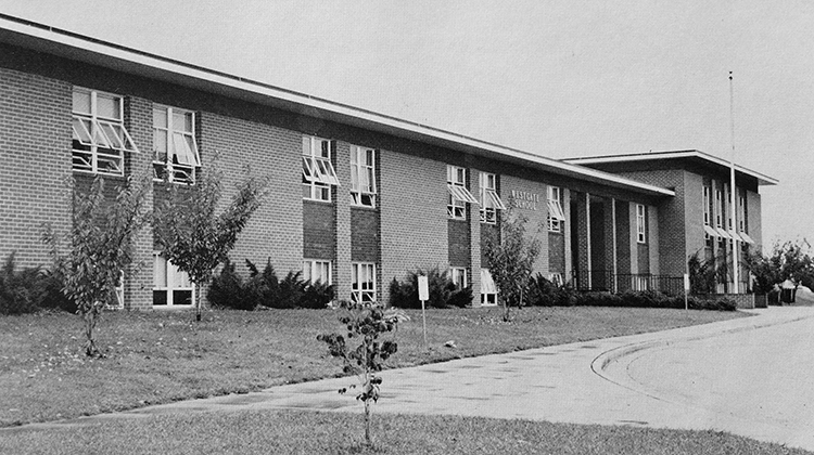Black and white photograph of the front of Westgate Elementary School from the 1974 to 1975 yearbook. The main entrance is visible on the far right. Given their size, the cherry trees that once graced the grounds in front of the building appear to have been planted relatively recently, perhaps two or three years prior. It is a warm day as all the windows are propped open. The building had no air-conditioning at this time.   