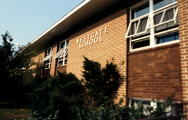 Color photograph of the front of Westgate Elementary School taken in the early 1980s. The name of the school is visible on the building. The classroom windows are all propped open because the building is still without air-conditioning.  